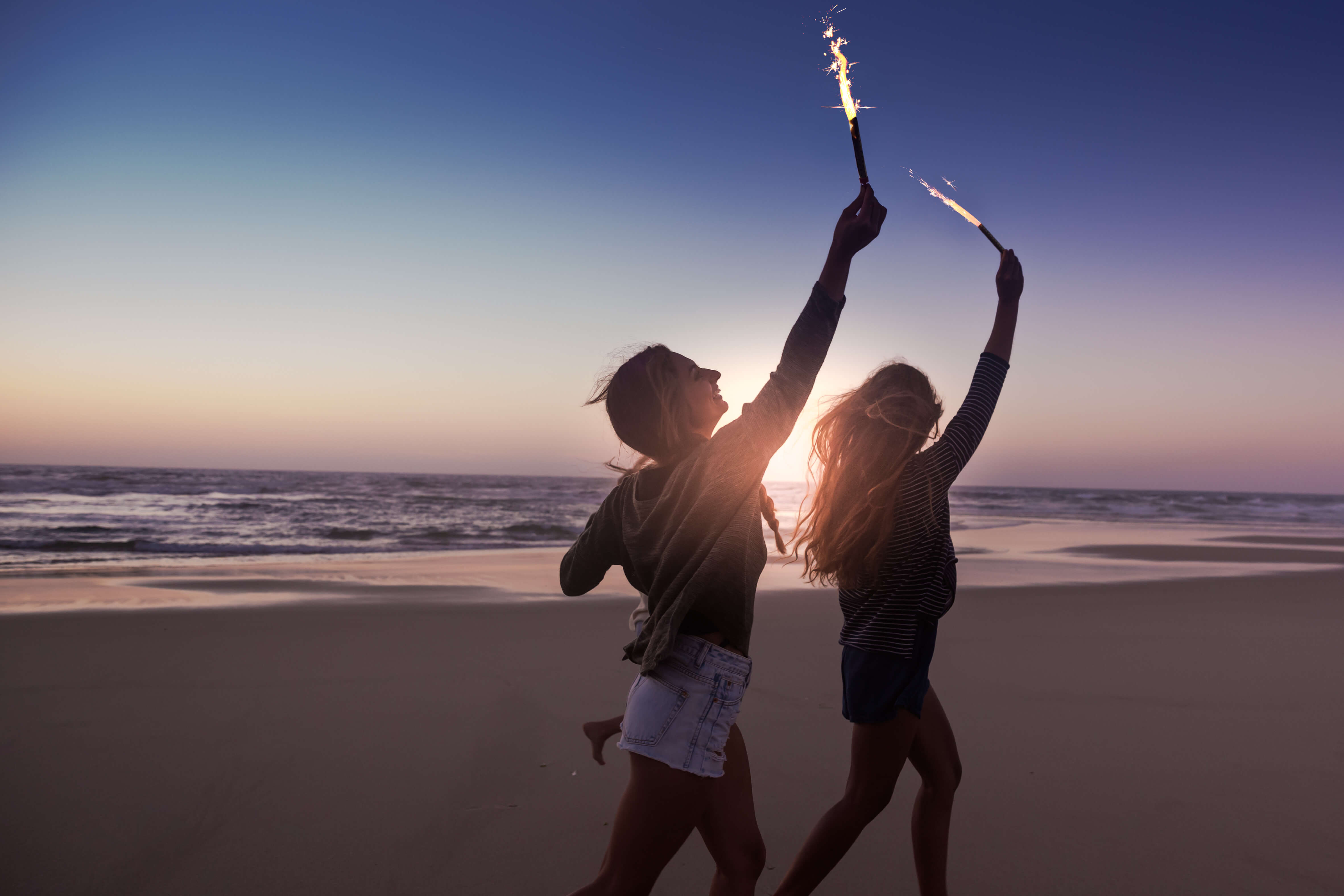 Girls smiling and running on the beach with sparklers while the sun sets over the ocean