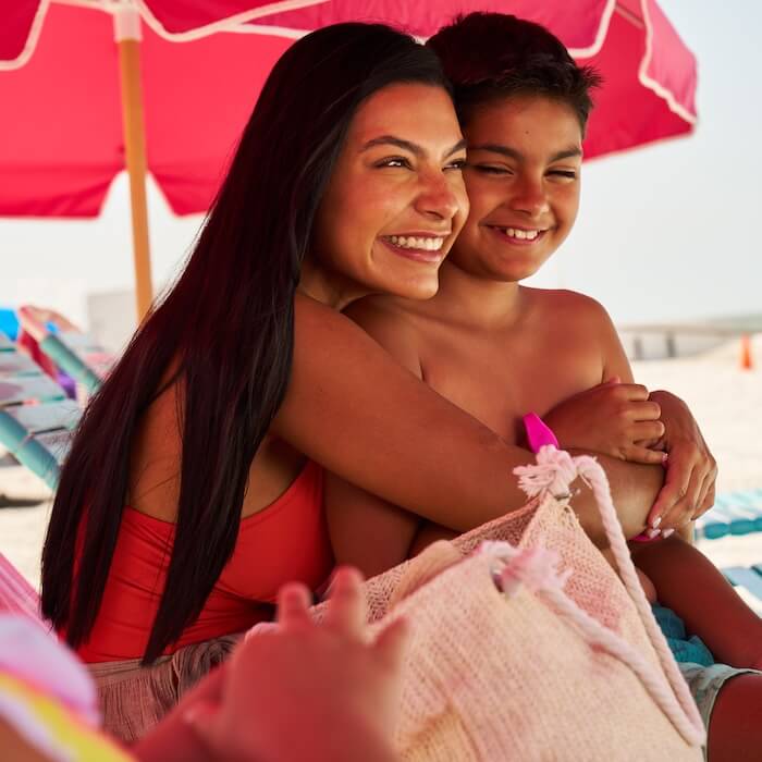 A mother and son hug and smile on a chair under an umbrella on Fort Myers Beach at Pink Shell Resort