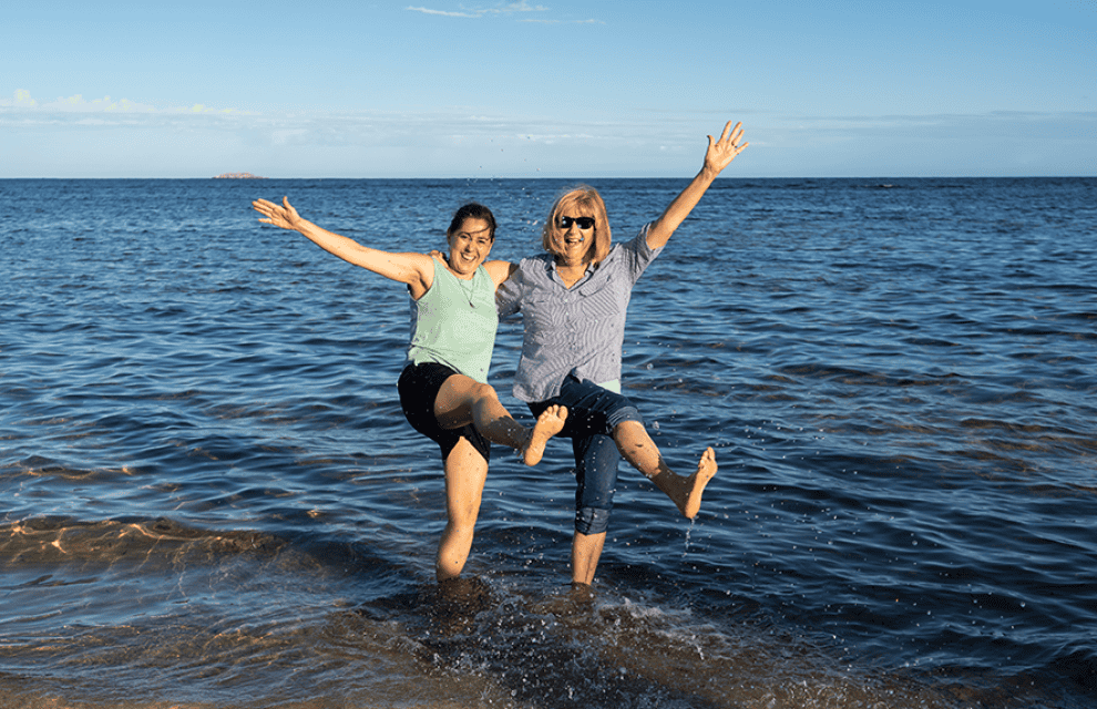 Two women stand in shallow ocean water, smiling and raising their arms and legs joyfully, with a clear sky in the background.