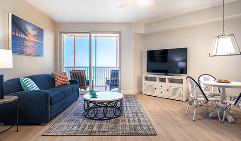 Modern living room with blue sofa, coffee table, TV, and dining area; large windows open to a balcony with ocean view.