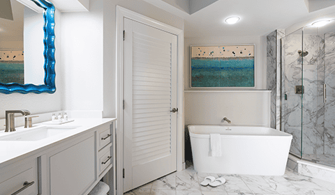 Modern bathroom with white vanity, freestanding bathtub, glass shower, marble tile floor, and a blue-framed mirror next to a colorful abstract painting above the tub.