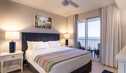 A neatly made bed with a striped blanket in a bright hotel room featuring lamps, artwork, and a balcony with an ocean view.