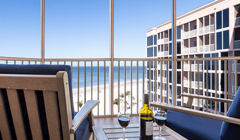 Two chairs on a balcony overlook a beach and ocean, with a table holding a bottle of wine and two glasses; a neighboring building is visible on the right.