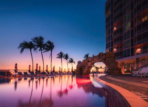 Outdoor pool next to a modern hotel at sunset, with palm trees, poolside lounge chairs, and colorful lights reflecting on the water.