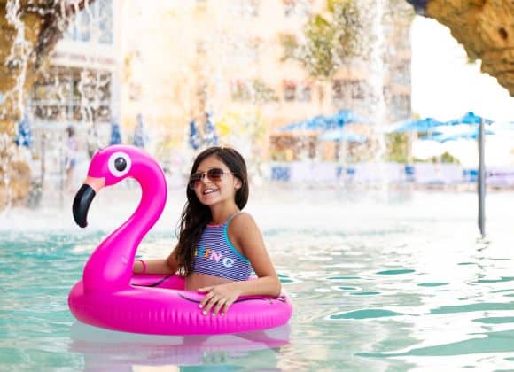 A girl in sunglasses sits in a pool using a pink inflatable flamingo float, with a waterfall and buildings in the background.
