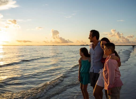 A family of four stands together on a beach at sunset, looking toward the horizon as gentle waves touch the shore.
