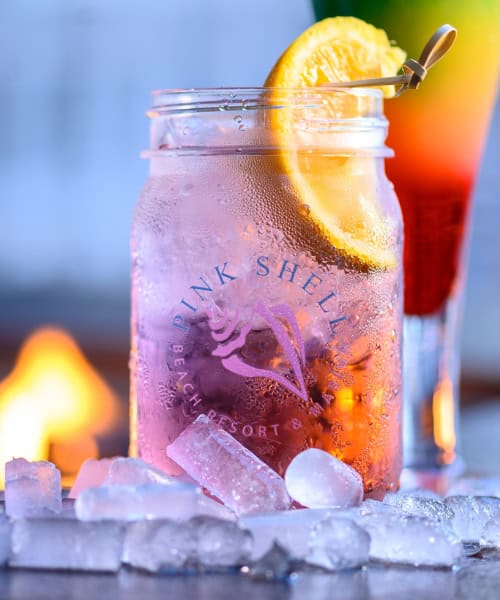 A mason jar with a Pink Shell Beach Resort logo, filled with a pink drink, ice, and a lemon slice, sits among ice cubes with a blurred drink and fire in the background.