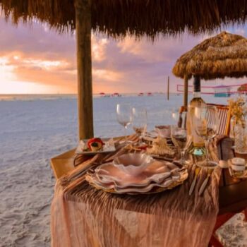 A table set for a meal with elegant plates and glassware sits under a thatched umbrella on a sandy beach at sunset.