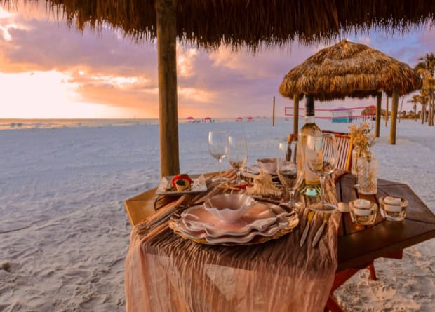 A table set for a meal with elegant plates and glassware sits under a thatched umbrella on a sandy beach at sunset.
