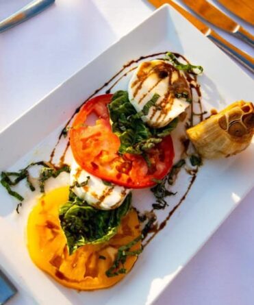 A Caprese salad with sliced tomatoes, mozzarella, basil, and balsamic glaze on a rectangular white plate, served with roasted garlic.