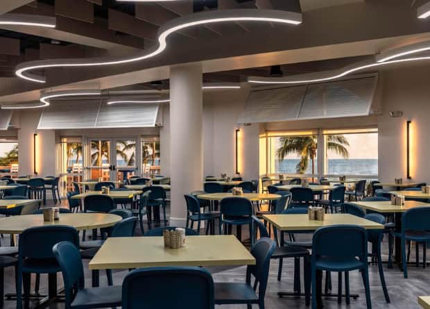 Modern dining area with yellow tables and blue chairs, large windows showing a view of palm trees and the ocean, and wavy ceiling lights.
