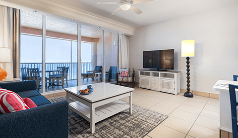 Modern living room with a white coffee table, dark blue sofa, TV on a stand, floor lamp, and a balcony with outdoor seating visible through large glass doors.