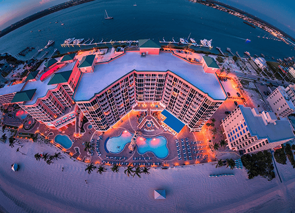 Aerial view of a large beachfront resort with illuminated pools at dusk, facing a calm bay with boats and a shoreline in the background.