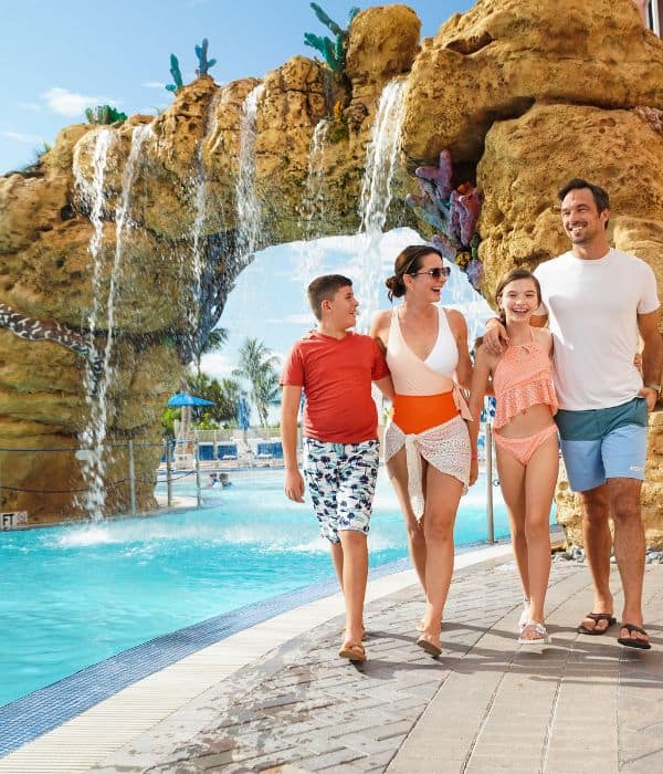 A family of four in swimwear walks together by a pool with a rocky waterfall feature in the background.