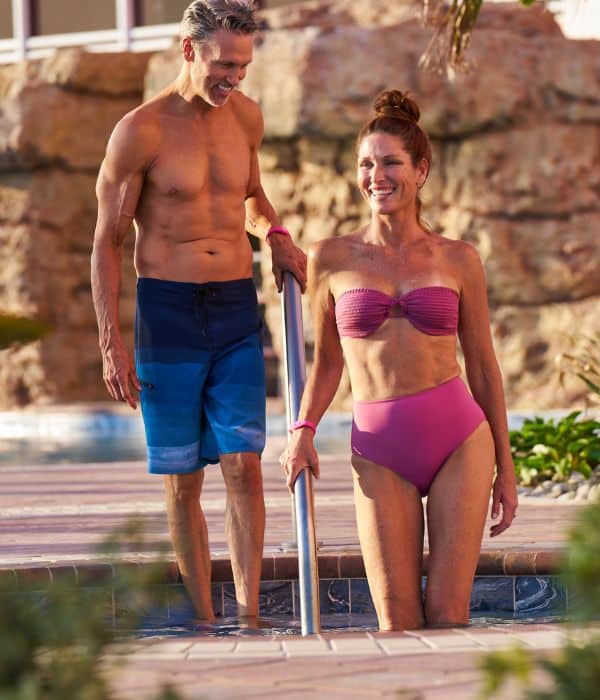 A man in swim trunks stands at the edge of a pool while a woman in a bikini steps into the water, both smiling outdoors near a rocky background.