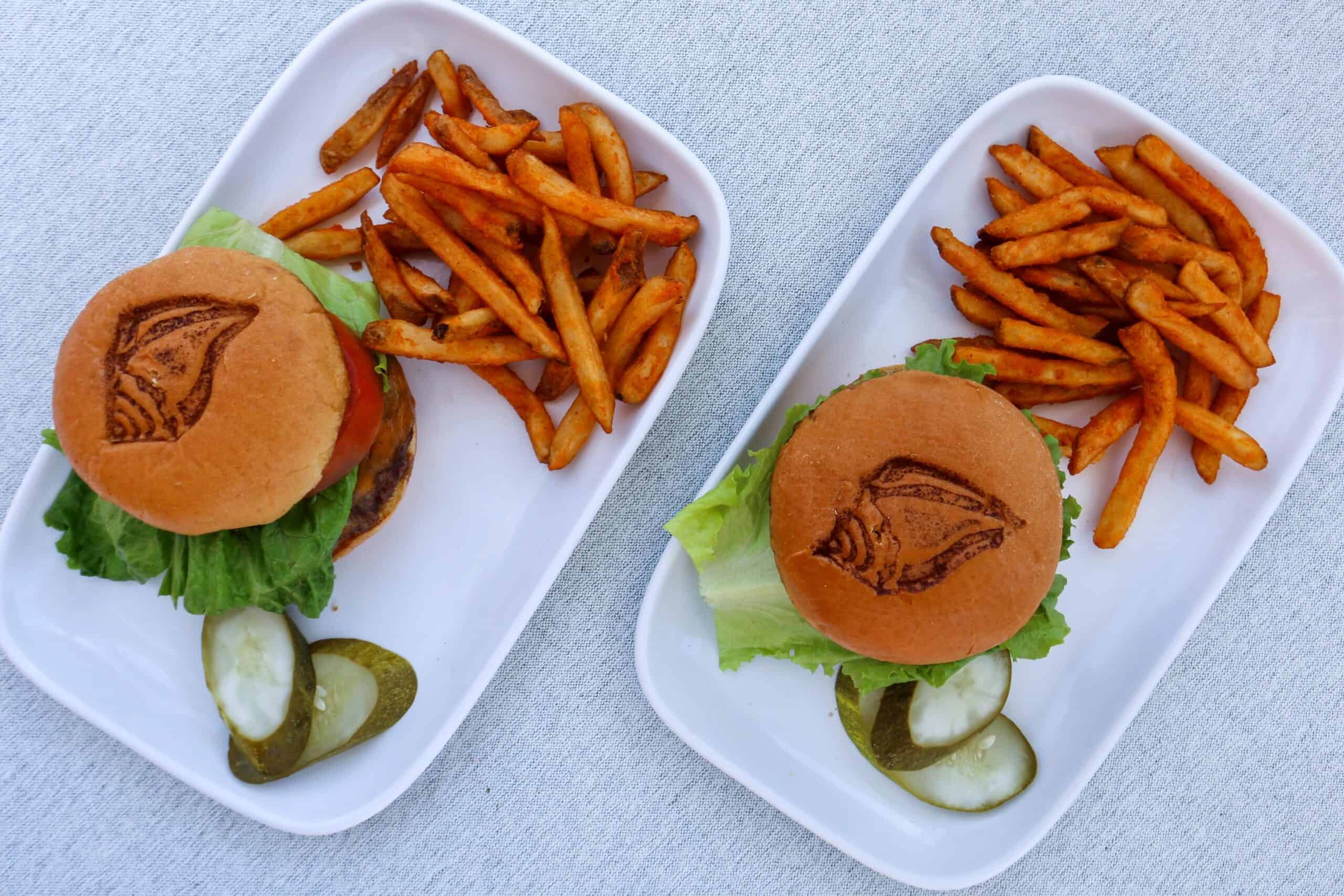 Two white plates each with a hamburger, French fries, and pickles. The hamburger buns have a Pink Shell logo toasted on the top. Plates rest on a light-colored tablecloth.