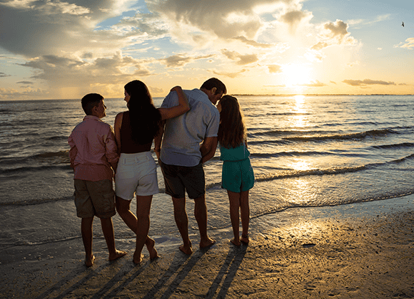 A family of four stands barefoot on a beach at sunset, looking out over the ocean with clouds and sunlight in the background.