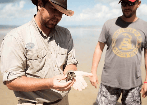A man in outdoor clothing holds a small crab in his hand while another man stands nearby on a sandy beach.