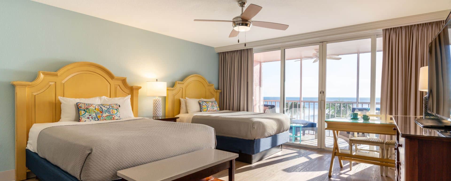 Hotel room with two double beds, a ceiling fan, desk, TV, and large windows opening to a balcony with ocean view.