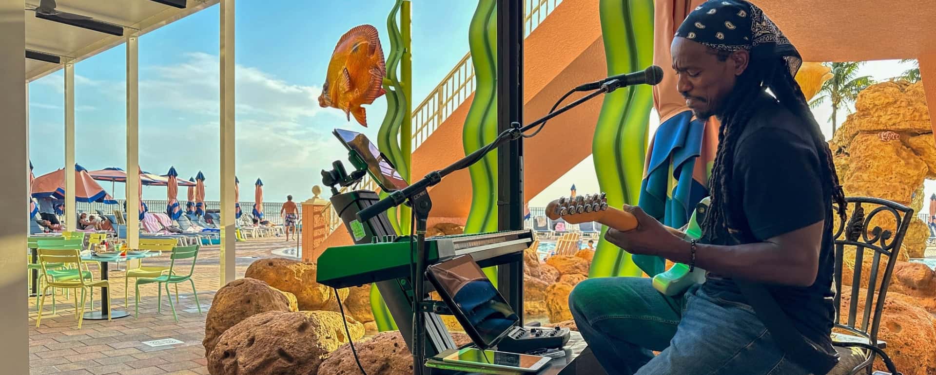 A musician sits outdoors playing an electric guitar and singing into a microphone, with poolside tables, umbrellas, and decorative elements in the background.