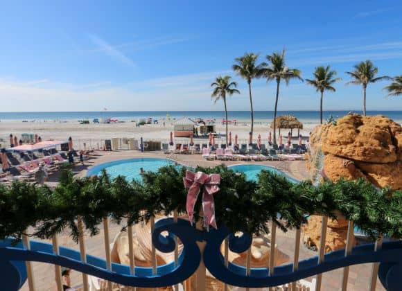A decorated balcony overlooks a pool, palm trees, and a sandy beach under a clear blue sky.