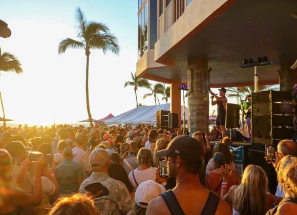 A crowd gathers outdoors near a stage at sunset, facing a performer. Palm trees and a tent are visible, with people taking photos and enjoying the event.