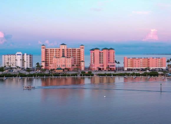 Three pastel-colored buildings line the waterfront at sunset, with a small boat moving across the calm water in the foreground.