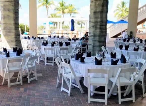 Outdoor event setup with round tables covered in white cloths, white chairs, and neatly folded dark napkins, surrounded by palm trees and a partly sunny sky.