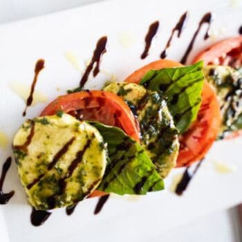 Slices of fresh mozzarella, tomato, and basil are arranged on a plate, topped with pesto and drizzled with balsamic glaze and olive oil.