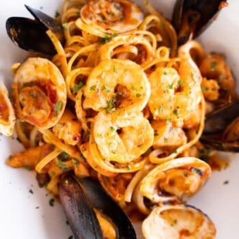 A plate of linguine pasta topped with clams, mussels, shrimp, and a tomato-based sauce, garnished with chopped herbs.