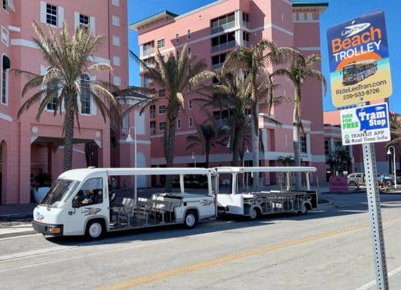 A white beach trolley with open sides is parked on a street near pink buildings and palm trees; a sign indicates a tram stop for the LeeTran Beach Trolley.