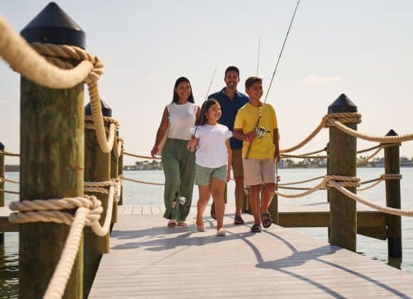 A family of four, holding fishing rods, walks on a wooden dock by the water on a sunny day.