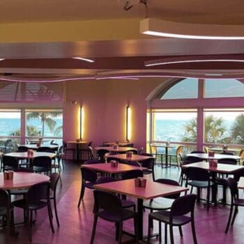 A modern dining area with purple lighting, empty tables and chairs, large windows, and a view of the ocean and palm trees outside.