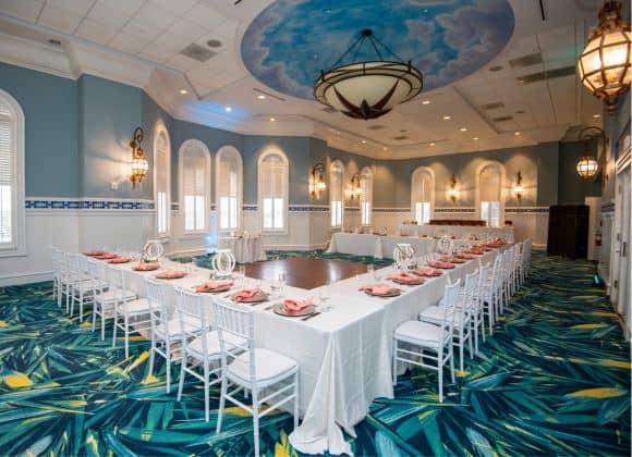 A banquet room with a U-shaped table set for a meal, white chairs, pink napkins, tropical-patterned carpet, and a painted ceiling with a sky design.