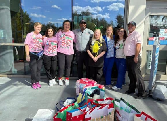 A group of eight adults stands outside a building, smiling at the camera, with several colorful gift bags placed on the ground in front of them.