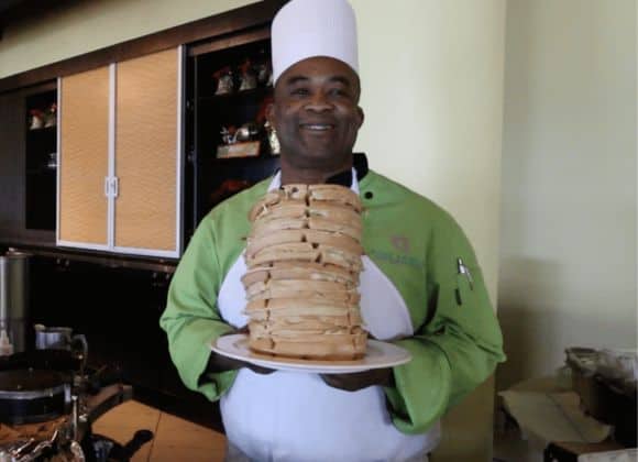 A chef in a green and white uniform holds a large plate stacked high with pancakes, smiling at the camera.
