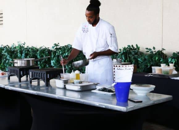 A chef in a white uniform prepares food at an outdoor cooking station with various pots, pans, and ingredients on the table.
