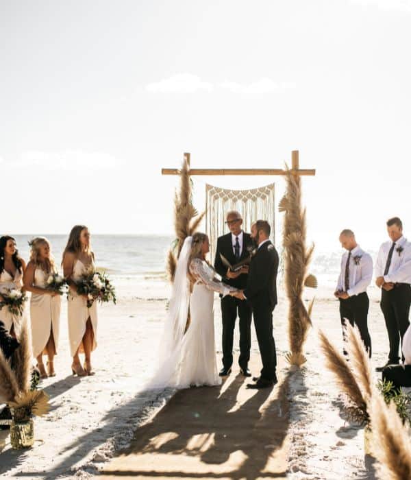 A wedding ceremony taking place on a beach with a couple holding hands in front of an officiant, surrounded by bridesmaids and groomsmen.