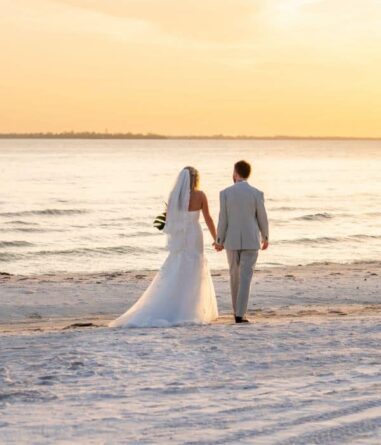 A bride and groom, dressed in wedding attire, walk hand in hand along a sandy beach at sunset, facing the ocean.