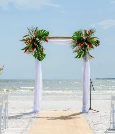 A bamboo wedding arch decorated with tropical leaves and flowers stands on a sandy beach, with white chairs and the ocean in the background.