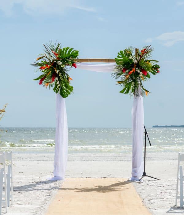 A bamboo wedding arch decorated with tropical leaves and flowers stands on a sandy beach, with white chairs and the ocean in the background.