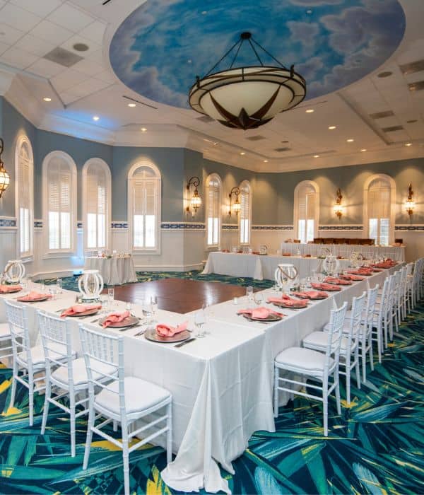 A banquet hall with U-shaped tables set for an event, white chairs, peach napkins, and a painted sky ceiling above a central dance floor.