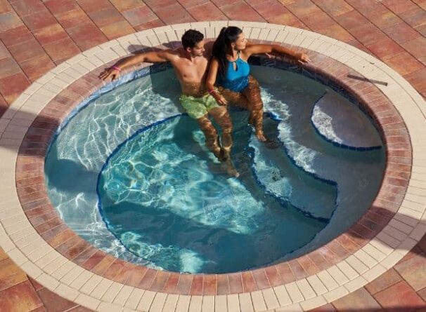 A man and a woman sit together in a circular outdoor hot tub with clear water, surrounded by a tile deck.
