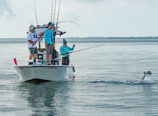 Three people fishing from a small boat in calm waters, with one person reeling in a large fish that is leaping out of the water near the boat.
