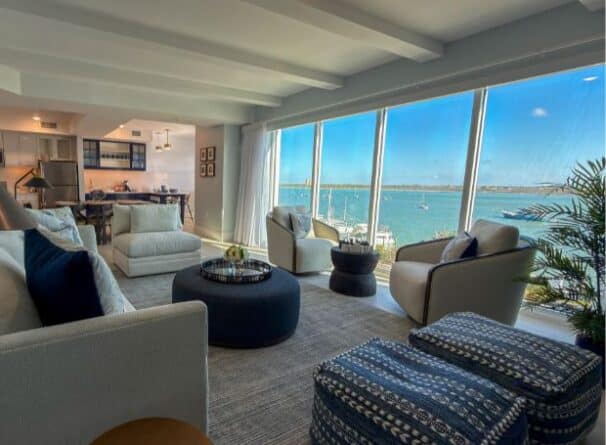 A modern living room with large windows overlooks a marina with boats. The space features neutral-toned furniture, a round ottoman, and a view of blue water and sky.
