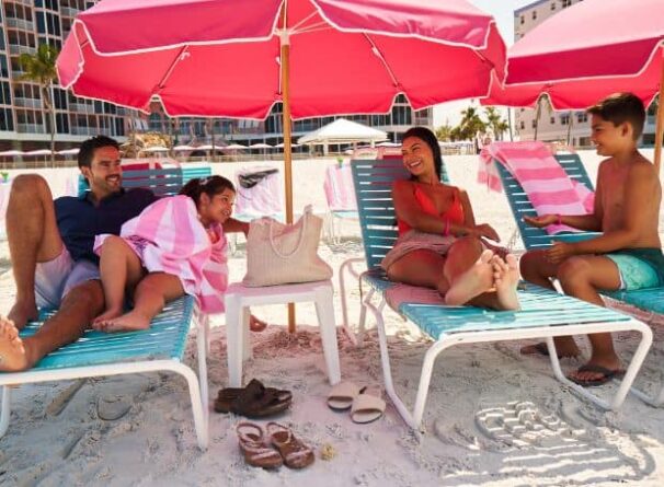 A family of four relaxes on lounge chairs under pink umbrellas at a sandy beach, with towels, sandals, and a bag nearby.