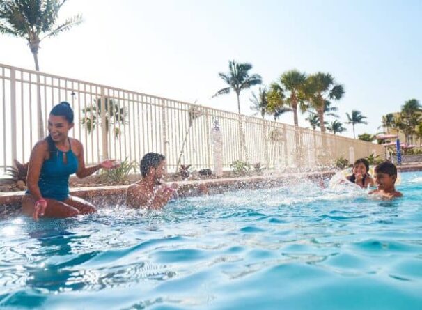 Four people are playing and splashing water in an outdoor swimming pool on a sunny day, with palm trees and a fence in the background.