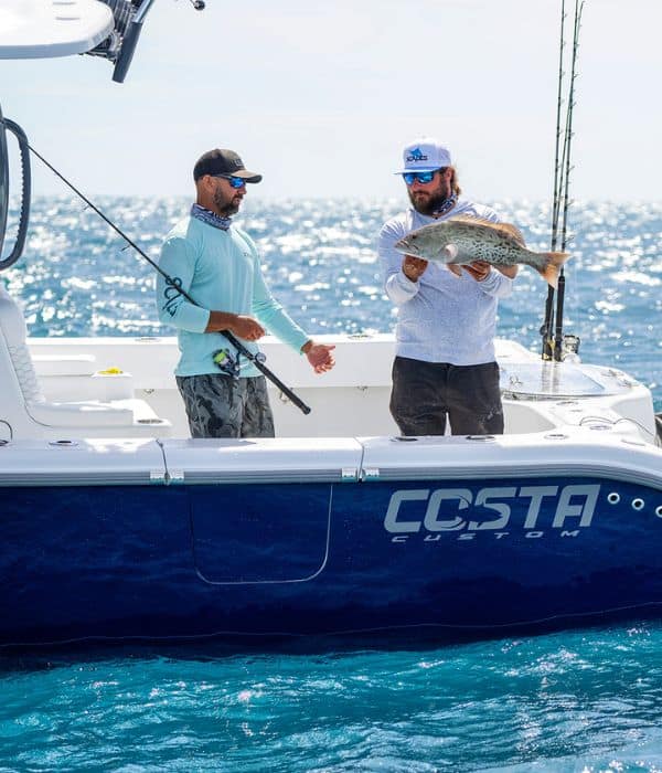 Two men on a fishing boat at sea; one holds a large fish while the other gestures beside him. The boat has 