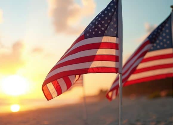 American flags on poles are displayed on a sandy beach at sunset, with the sky and sun creating a warm, glowing background.