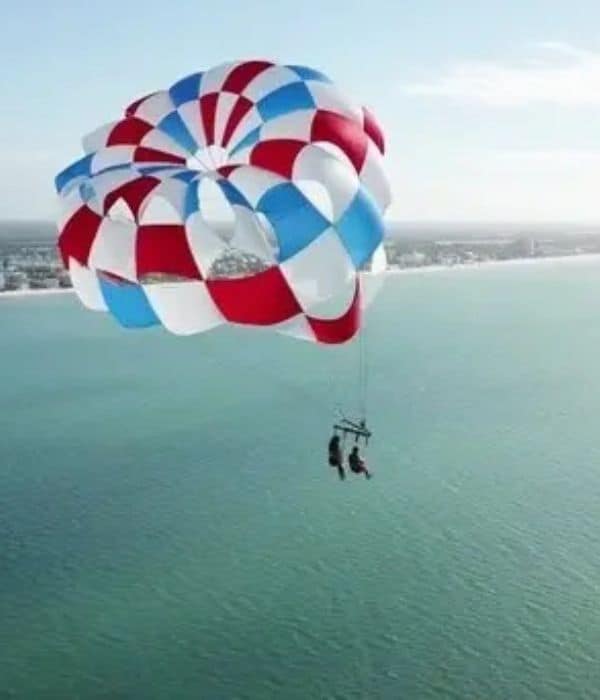 Two people parasailing over the ocean with a red, white, and blue parachute, with a coastline visible in the background.